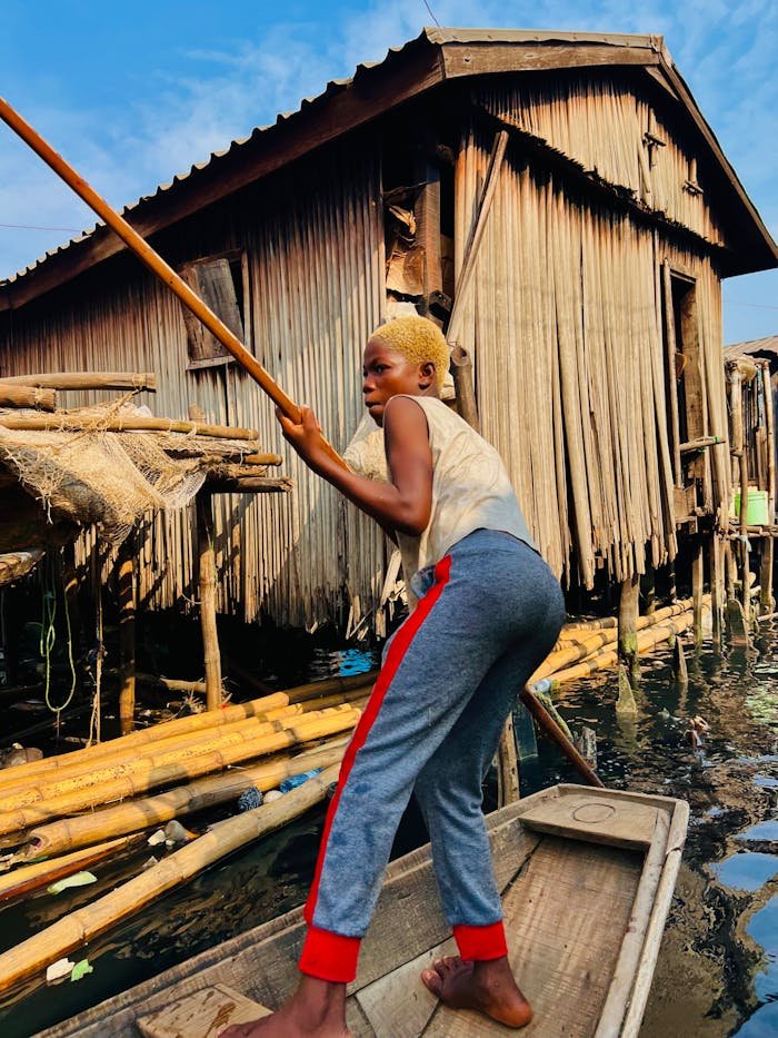 A woman skillfully paddles a small wooden boat through a rural waterway village, surrounded by wooden structures.
