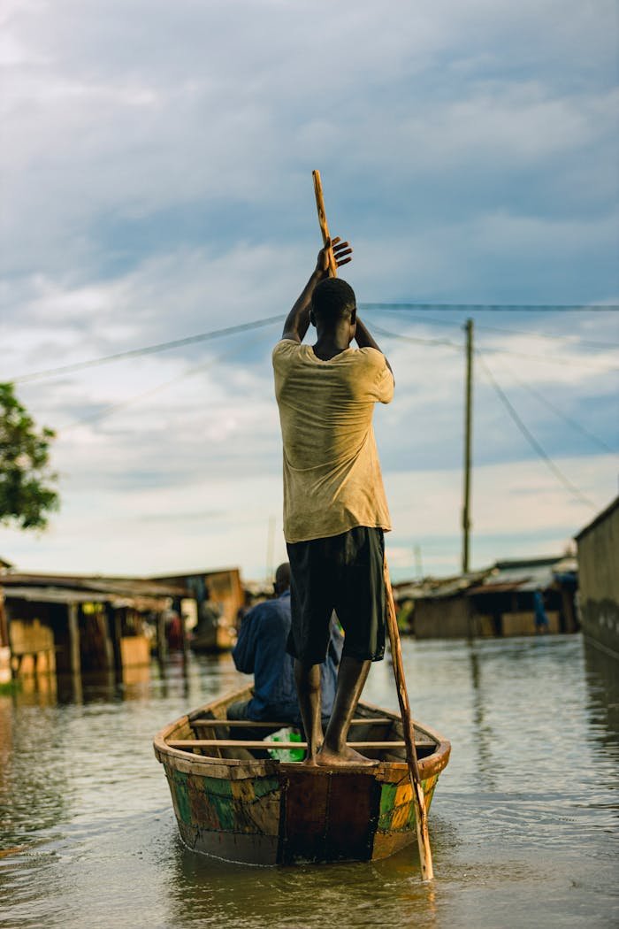 A man stands in a wooden boat, paddling through a flooded rural village.