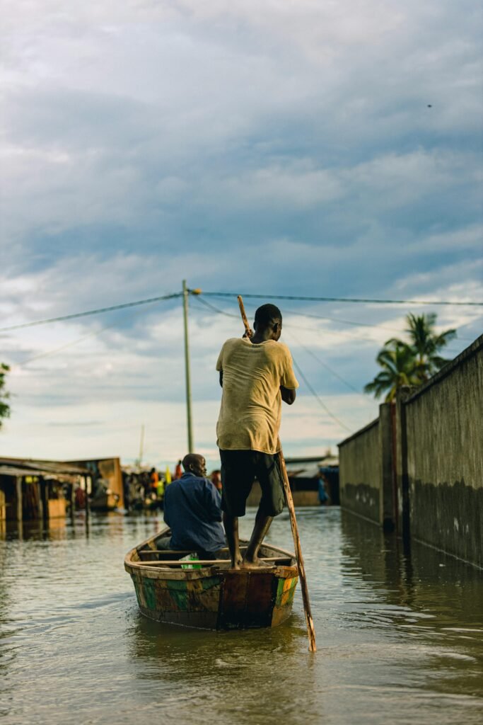 Environmental Justice Two men navigate a wooden boat through a tranquil canal setting under a cloudy sky.